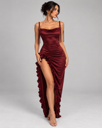 Femme portant la Robe Nina bordeaux satinée, vue de face, avec fines bretelles, encolure drapée, coupe longue ajustée et fente haute bordée de volants, dans un style glamour et sensuel