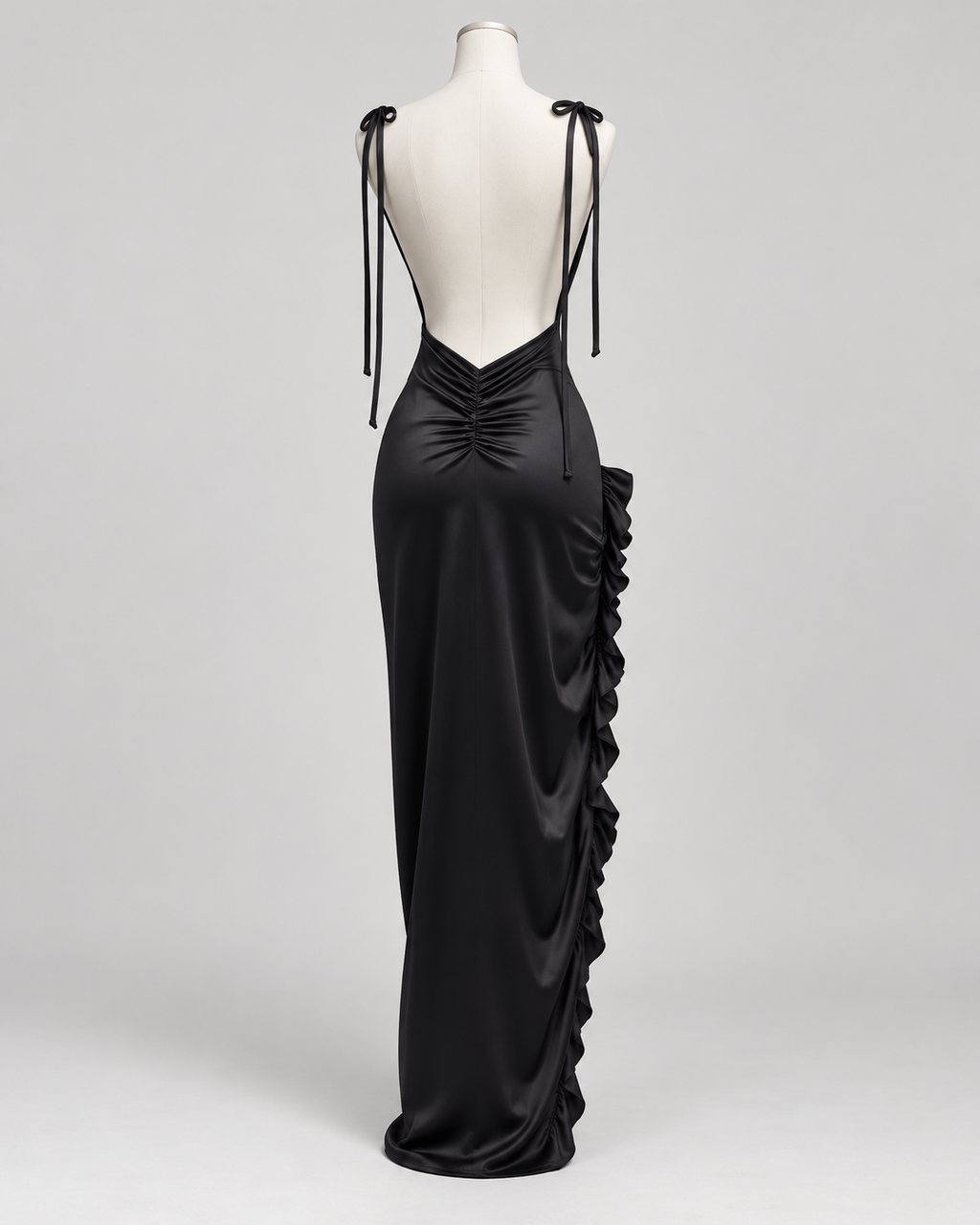 Robe Nina noire satinée présentée sur mannequin, vue de dos, avec dos nu plongeant, fines bretelles à nouer, fronces au bas du dos et volants sur le côté